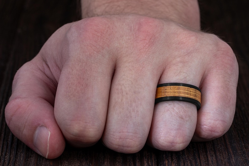 May include: A ring featuring a matte black band with a central inlay of light brown wood. The wood inlay is divided into three horizontal sections by two thin black lines, creating a layered appearance around the circumference of the ring.
