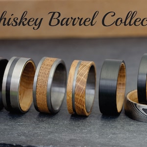 May include: A collection of six men's wedding bands featuring a combination of black and silver metal with wood inlays. The rings are arranged in a row on a dark gray surface. "Whiskey Barrel Collection" is written in black cursive above the rings.