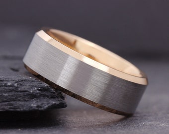 May include: A close-up of a two-tone ring. The ring features a brushed silver band with a gold-colored interior and edges. The ring is set against a dark, textured background, highlighting its metallic sheen.