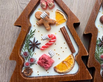 Pode incluir: Uma vela em forma de árvore de Natal com moldura de madeira. A vela apresenta uma base de cera branca com elementos decorativos, incluindo um homem de gengibre, um bastão de doces, uma fatia de laranja e um pau de canela. Um laço rosa e bagas vermelhas também estão incluídos.