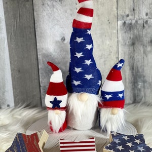 May include: Three gnomes wearing red, white, and blue hats with stars and stripes. The gnomes are standing on a white surface with three wooden stars with red, white, and blue designs.