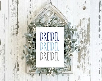 May include: White sign with the word "Dreidel" written three times in blue, light blue, and gray lettering. The sign is framed by a white wreath with green leaves and white berries.