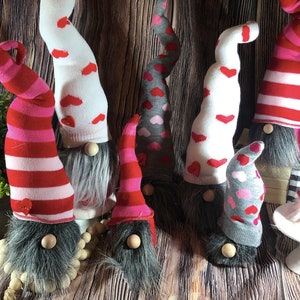 May include: A collection of nine Valentine's Day gnomes with red, pink, white, and gray striped and heart patterned hats. The gnomes are made from fabric and have fuzzy gray beards.