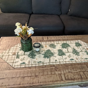 May include: A beige and green table runner with a woodland print featuring trees and leaves. The runner is on a wooden table with a vase of flowers and a candle holder.