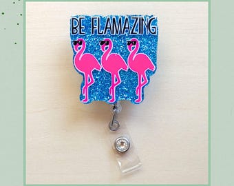 May include: A retractable badge reel with a blue glitter background and the text "BE FLAMAZING". The badge reel features three pink flamingos wearing sunglasses. The reel has a clear plastic strap and a silver-colored metal clip.