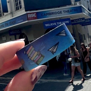 Op de afbeelding: Een blauw en wit armband met een papieren vliegtuig en een ontwerp van gestapelde boeken. Het armbandje zegt "Believe the Hype" en bevindt zich voor het Cambridge Theatre, dat reclame maakt voor de musical "Matilda".