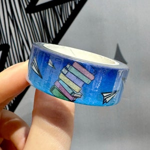 Op de afbeelding: Een blauwe en witte washi tape met een stapel kleurrijke boeken en papieren vliegtuigjes.