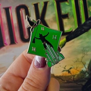 May include: Green enamel pin featuring a silhouette of a witch flying with a broom over a green city. The pin is in front of a book cover with the word "WICKED" in large gold letters.