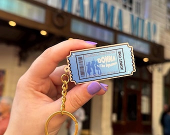 May include: A light blue ticket-shaped keychain featuring a gold-tone metal chain and ring. The ticket design includes the text 'DONNA and The Dynamos', 'ADMIT ONE', a barcode, and silhouettes of three figures, accented with small star graphics.