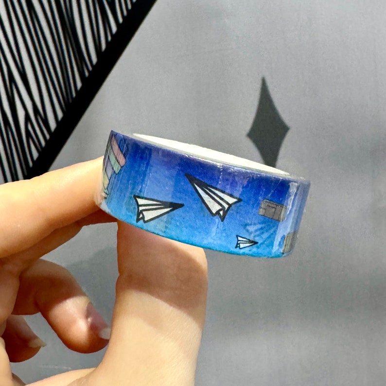 Op de afbeelding: Een rol blauwe washi tape met een verloopontwerp met witte papieren vliegtuigjes en grijze rechthoekige vormen.