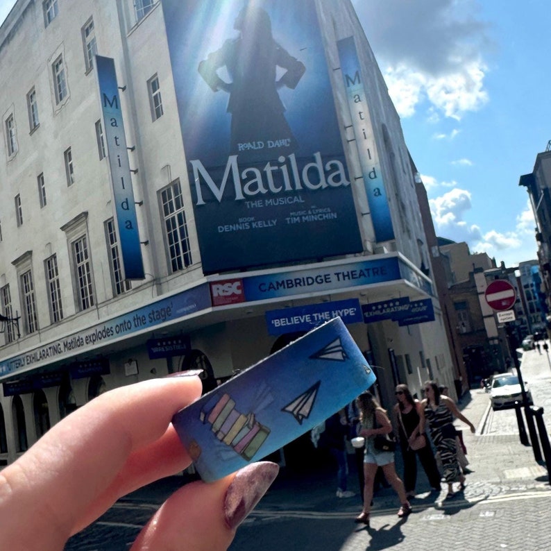 Op de afbeelding: Een blauw en wit armband met een papieren vliegtuig en een ontwerp van gestapelde boeken. Het armbandje zegt "Believe the Hype" en bevindt zich voor het Cambridge Theatre, dat reclame maakt voor de musical "Matilda".