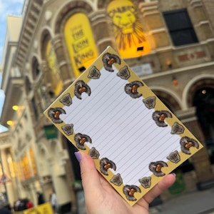 May include: A lined notepad with a yellow border decorated with lion faces. The notepad is held in front of a building with a sign that says "The Lion King". The notepad has a white background.