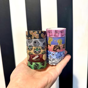 May include: A collection of eight colorful washi tapes featuring various designs, including a gingerbread man, a Star Wars character, a green turtle, a leaf, a cat, and a game controller. The tapes are stacked in two columns and held in a hand.