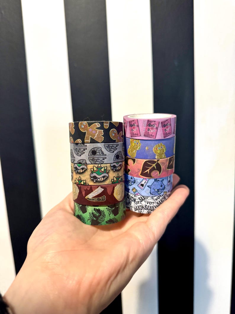 May include: A collection of eight colorful washi tapes featuring various designs, including a gingerbread man, a Star Wars character, a green turtle, a leaf, a cat, and a game controller. The tapes are stacked in two columns and held in a hand.