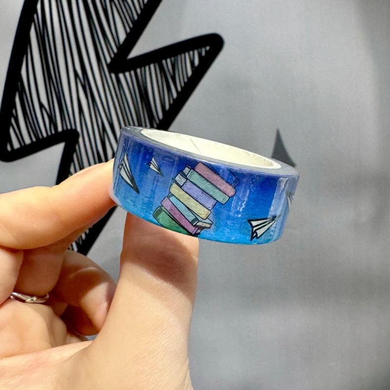 Op de afbeelding: Een blauwe en witte washi tape met een stapel kleurrijke boeken en papieren vliegtuigjes.