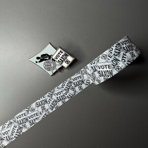 May include: A white and black patterned washi tape with the text "Vote Saxon" repeated on the tape. A small metal pin with a black silhouette of a person holding a sign that says "Vote Saxon" is also in the image.
