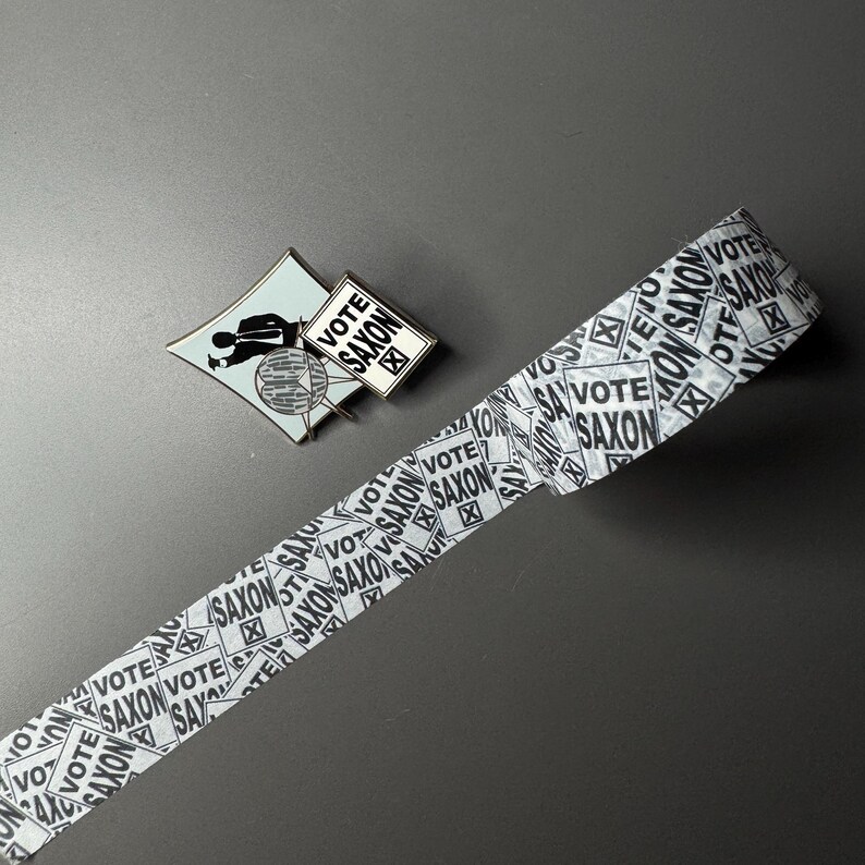 May include: A white and black patterned washi tape with the text "Vote Saxon" repeated on the tape. A small metal pin with a black silhouette of a person holding a sign that says "Vote Saxon" is also in the image.
