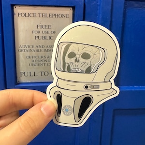 Könnte beinhalten: Weißer Astronautenhelm-Aufkleber mit einem schwarzen Schädel in der Visiere des Helms. Der Aufkleber ist teilweise von einer blauen Oberfläche abgezogen. Der Text "PULL TO" ist auf der Oberfläche sichtbar.