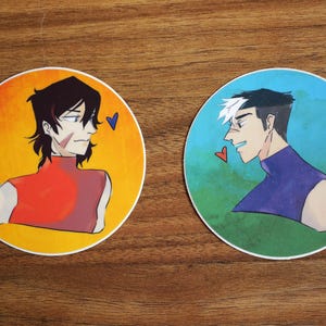 Peut inclure: Deux autocollants ronds avec des illustrations de deux hommes en dessin anim&eacute;. L'homme &agrave; gauche a les cheveux noirs et porte une chemise rouge. L'homme &agrave; droite a les cheveux blancs et porte une chemise violette. Les deux autocollants ont une forme de c&oelig;ur.