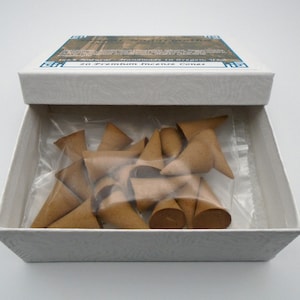 Puede incluir: Una caja blanca con una etiqueta azul y blanca que dice "20 Premium Incense Cones" y "Incense Handmade in Oregon, USA". La caja est&aacute; abierta y contiene una bolsa de pl&aacute;stico transparente llena de 20 conos de incienso marrones.