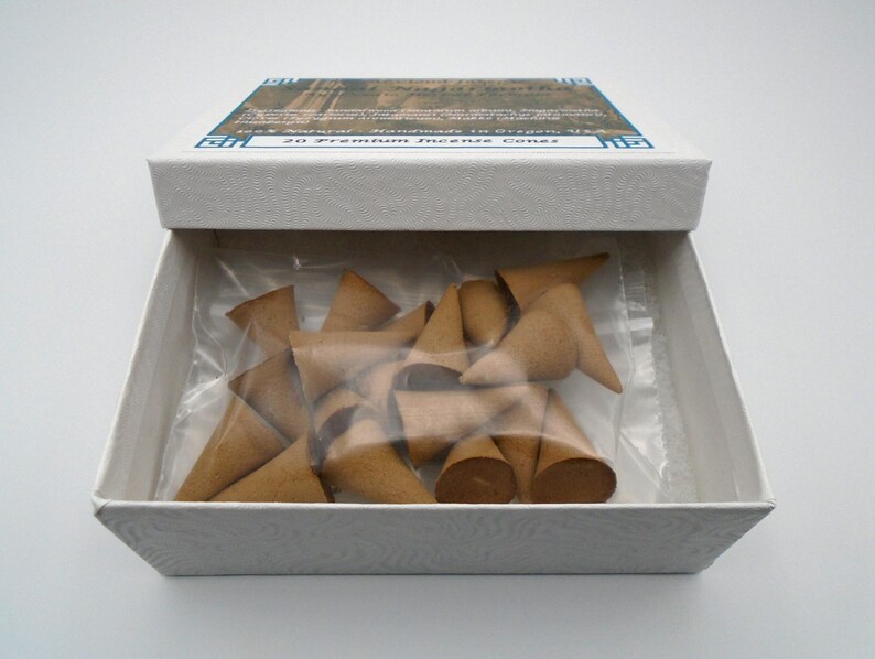 Puede incluir: Una caja blanca con una etiqueta azul y blanca que dice "20 Premium Incense Cones" y "Incense Handmade in Oregon, USA". La caja est&aacute; abierta y contiene una bolsa de pl&aacute;stico transparente llena de 20 conos de incienso marrones.