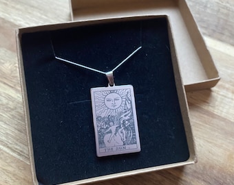May include: A silver-toned pendant necklace featuring a rectangular charm with an engraved tarot card design. The card depicts a sun with a face, figures, and the text "THE SUN." The necklace is presented in a brown box with a black interior.
