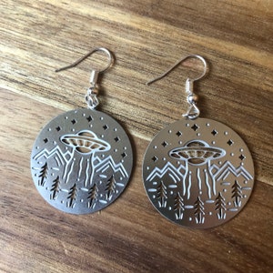 May include: Silver-toned metal earrings with a circular design. Each earring features a cut-out image of a UFO hovering over a mountain range and trees, with stars in the sky. The earrings have a hook closure.