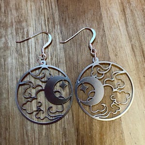 May include: Silver-toned earrings featuring a celestial design. Each earring has a circular frame with a crescent moon and two stars, surrounded by cloud-like cutouts. The earrings hang from curved ear wires.
