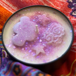 May include: A decorative candle in a black tin. The candle features a light purple floral design with glitter accents. The candle has a white base and a single wick. The background is a colorful fabric.