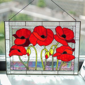 May include: Stained glass panel depicting red poppy flowers with yellow centres and green stems. The artwork is framed with a clear, textured border and hangs in front of a window. The panel is approximately 30 cm wide and 20 cm tall.
