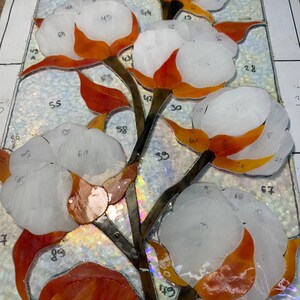 May include: A stained glass panel depicting a cotton plant. The artwork features white cotton bolls with orange and red accents, brown stems, and green leaves. The glass pieces are numbered and arranged on a white surface.