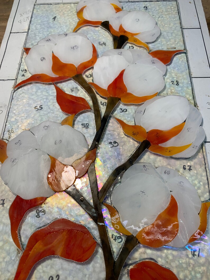 May include: A stained glass panel depicting a cotton plant. The artwork features white cotton bolls with orange and red accents, brown stems, and green leaves. The glass pieces are numbered and arranged on a white surface.