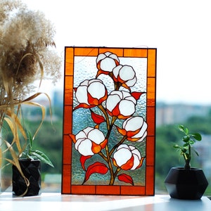 May include: Stained glass panel featuring a cotton plant design. The artwork showcases white cotton bolls with orange and red accents, set against a textured glass background. The panel is framed with an orange border, creating a decorative piece.