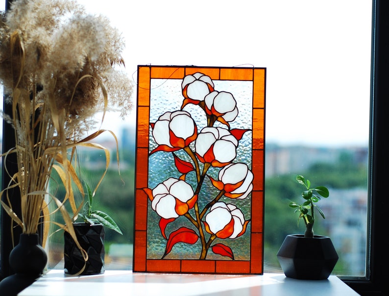 May include: Stained glass panel featuring a cotton plant design. The artwork showcases white cotton bolls with orange and red accents, set against a textured glass background. The panel is framed with an orange border, creating a decorative piece.