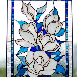 Puede incluir: Una ventana de vitrales que representa un grupo de flores blancas de magnolia con hojas y tallos azules. Las flores est&aacute;n dispuestas en forma de ramo y se encuentran sobre un fondo de cuadrados azules y blancos.