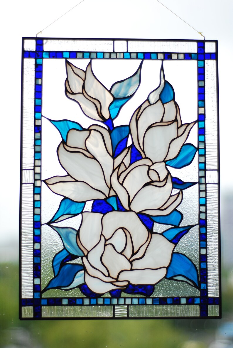 Puede incluir: Una ventana de vitrales que representa un grupo de flores blancas de magnolia con hojas y tallos azules. Las flores est&aacute;n dispuestas en forma de ramo y se encuentran sobre un fondo de cuadrados azules y blancos.