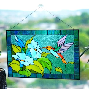 May include: Stained glass panel featuring a hummingbird and flowers. The hummingbird has orange, green, and purple wings, with a long beak reaching towards a light blue flower. The background is a textured teal color, framed by a dark blue border.