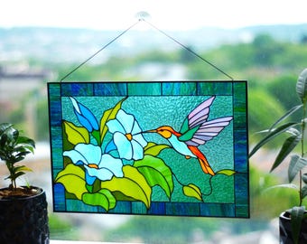 May include: Stained glass panel featuring a hummingbird and flowers. The hummingbird has orange, green, and purple wings, with a long beak reaching towards a light blue flower. The background is a textured teal color, framed by a dark blue border.