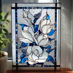 Puede incluir: Un panel de vitral con flores de magnolia blancas con hojas azules y blancas. El panel est&aacute; enmarcado en madera y tiene un fondo de vidrio transparente.