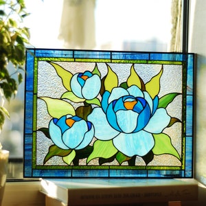 May include: A stained glass window panel featuring three blue and white flowers with green leaves. The panel is framed in blue and green glass.