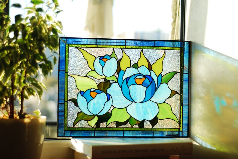 May include: A stained glass window panel featuring three blue and white flowers with green leaves. The panel is framed in blue and green glass.