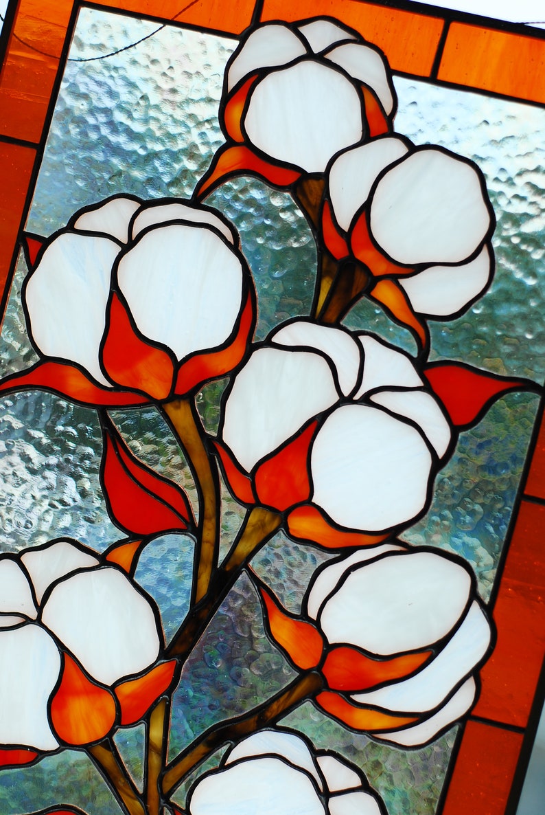 May include: Stained glass panel featuring cotton plants. The artwork showcases white cotton bolls with orange and red accents, set against a textured, light blue background. The panel is framed with an orange border.
