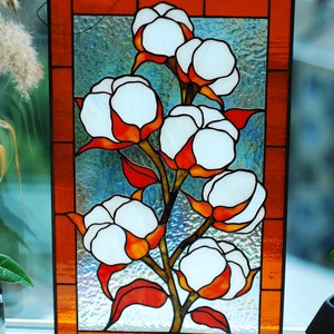 May include: Stained glass panel featuring a cotton plant design. The artwork showcases white cotton bolls with orange and red accents, set against a textured, light blue background. The panel is framed with an orange border.