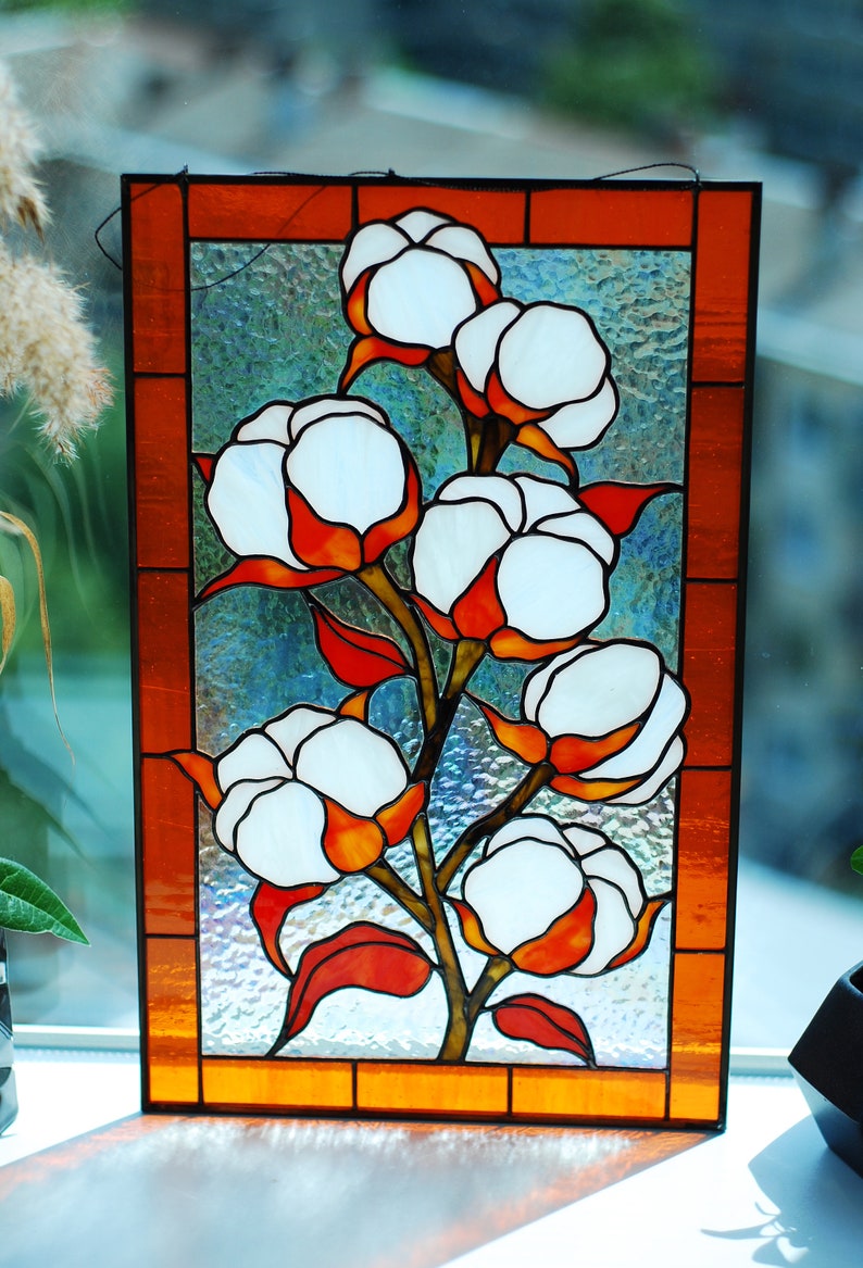 May include: Stained glass panel featuring a cotton plant design. The artwork showcases white cotton bolls with orange and red accents, set against a textured, light blue background. The panel is framed with an orange border.