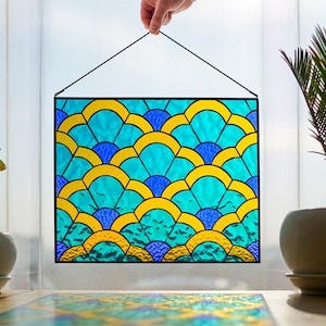 May include: A rectangular stained glass panel with a repeating pattern of turquoise, yellow, and blue arches. The panel is framed in black and suspended by a black cord. Two potted plants flank the panel.