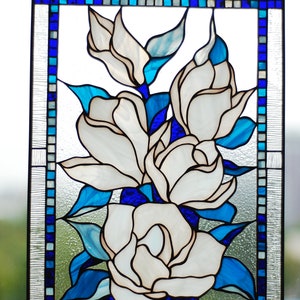 Puede incluir: Un panel de vitral con un ramo de flores blancas de magnolia con hojas azules. Las flores est&aacute;n dispuestas en un racimo y est&aacute;n rodeadas por un borde de cuadrados azules y blancos.