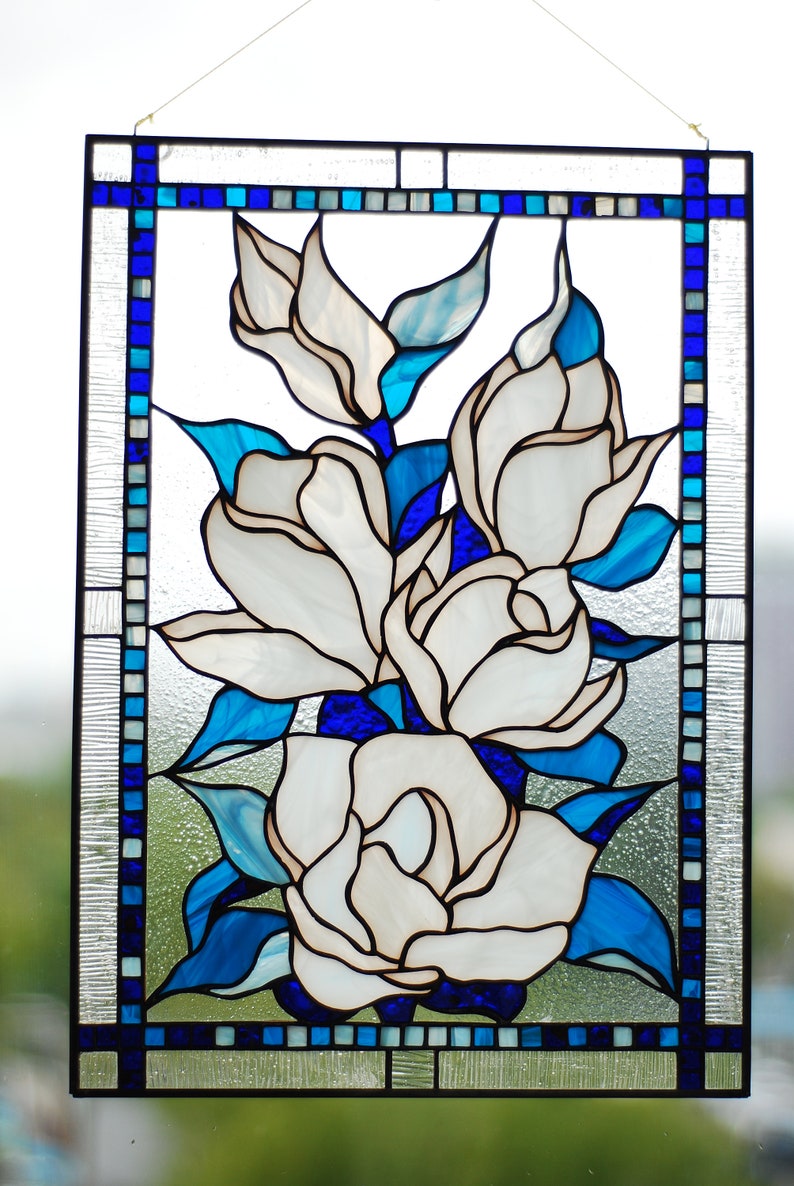 Puede incluir: Un panel de vitral con un ramo de flores blancas de magnolia con hojas azules. Las flores est&aacute;n dispuestas en un racimo y est&aacute;n rodeadas por un borde de cuadrados azules y blancos.