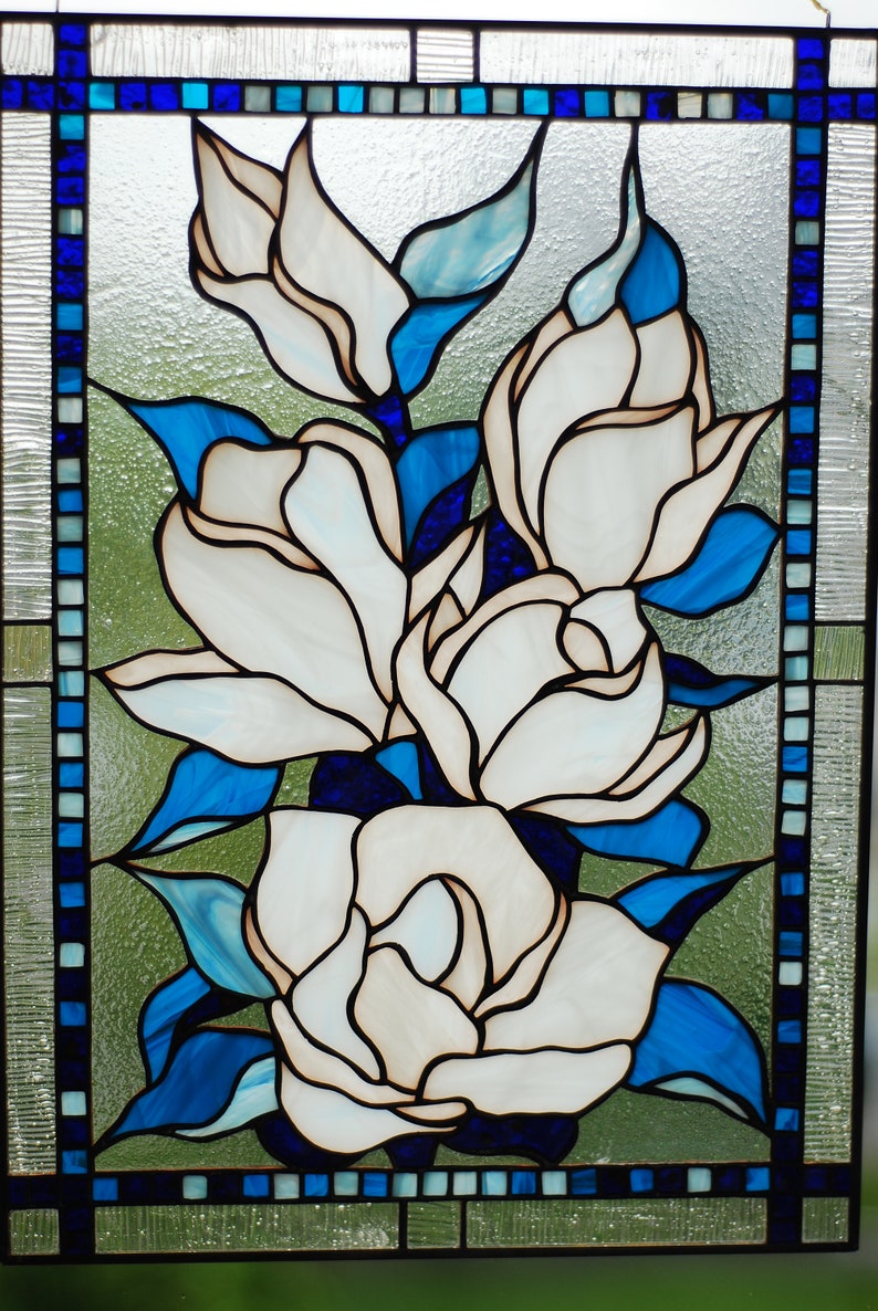 Puede incluir: Una ventana de vitrales que representa flores blancas de magnolia con hojas azules y un borde azul.
