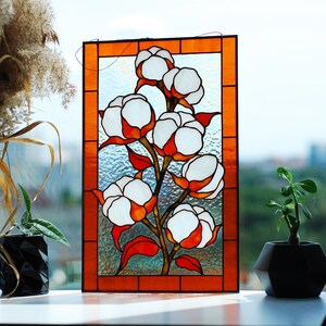 May include: Stained glass panel featuring a cotton plant design. The artwork showcases white cotton bolls with orange and red accents, set against a textured glass background. The panel is framed with an orange border, creating a decorative piece.