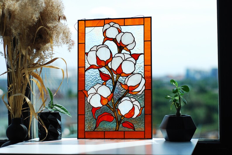May include: Stained glass panel featuring a cotton plant design. The artwork showcases white cotton bolls with orange and red accents, set against a textured glass background. The panel is framed with an orange border, creating a decorative piece.